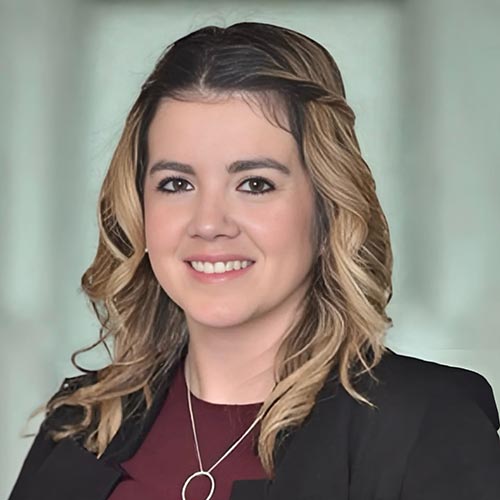 Alexandra Ridgeway; A woman with wavy blonde hair smiles confidently. She wears a black blazer over a burgundy top, with a necklace. The background is softly blurred.