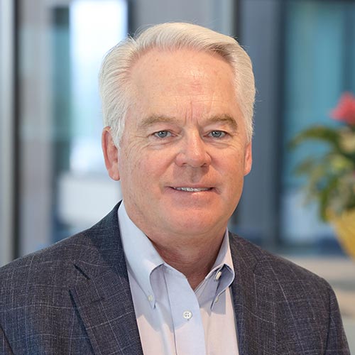 John Pike; A portrait of a white haired man wearing a light blue button up shirt and a grey blazer.