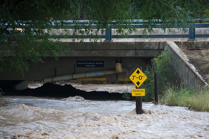 Flood Insurance Compliance in Colorado’s Mountain and Front Range Corridors; Floodwaters rush under a bridge marked "South Public Road" with a height restriction sign showing 7 feet. The scene conveys urgency and risk.