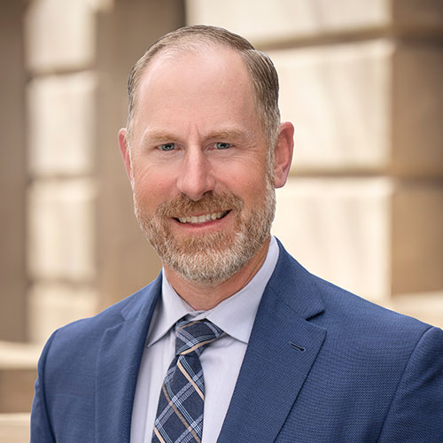 Kaycee Lytle; Man in a blue suit and patterned tie smiles warmly with a neutral background of blurred architectural elements, conveying professionalism and approachability.
