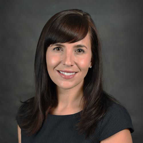 Natalie Strauss, a portrait of a brown haired women wearing a black and grey striped shirt, smiling with a black background