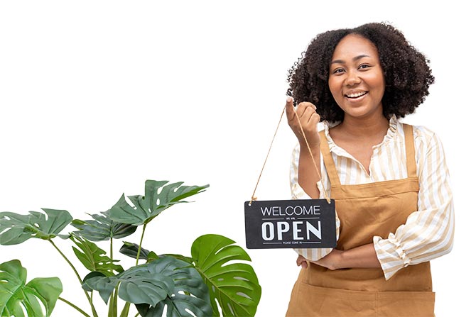 Why SBA Lending Holds Steady When the World Does Not; A woman in an apron smiles while holding a "Welcome, Open" sign. She stands next to green monstera plants, conveying a welcoming and cheerful ambiance.