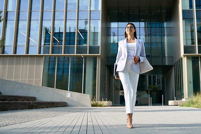 Women Leading the Way; A confident woman in a white suit strides purposefully in front of a modern glass building, carrying a bag and notebook, embodying professionalism and determination.