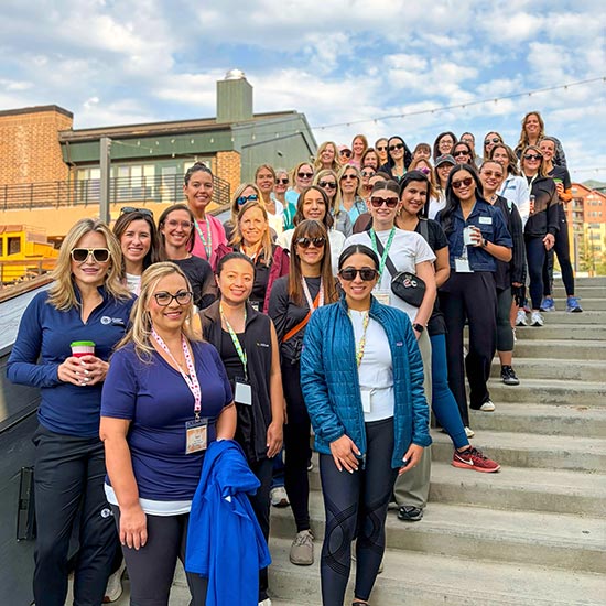 2026-Women-in-Banking-FEATURE - Large group of Women standing together on a set of steps outside all facing and smiling at the camera on a partly cloudy day.