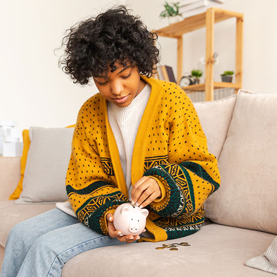 What-to-Know-FEATURE - Young African American woman placing coins into a piggy bank while seated on a couch.