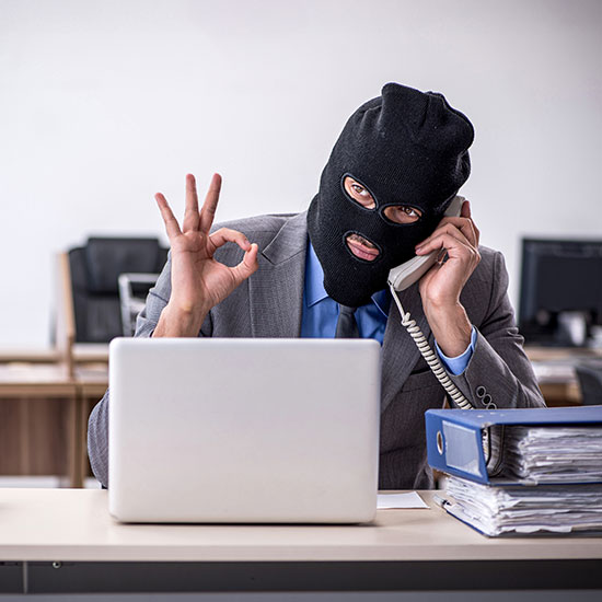 Why-Bank-Customers-FEATURE - Man in a business suit and a ski mask sitting at a computer and talking on a phone scamming someone.